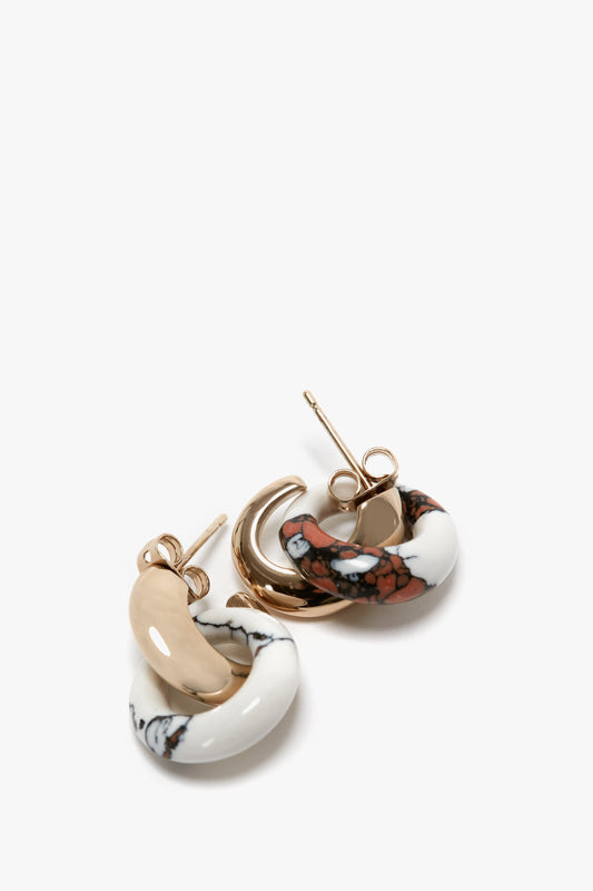 A set of three hoop earrings, crafted in Italy, are displayed on a white surface. The collection includes Victoria Beckham Exclusive Resin Pendant Earrings In Light Gold-White with black marbling and red with white and black patterns.