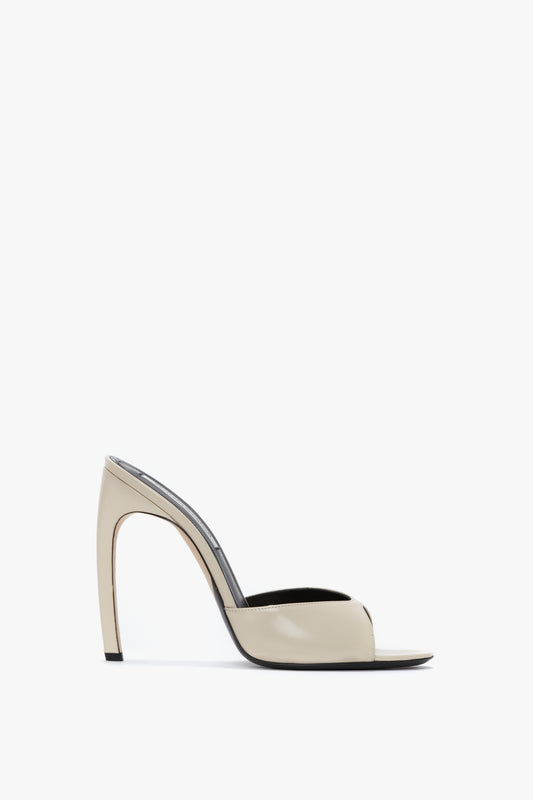 A side view of the Victoria Beckham Classic Mule In Macadamia Calf Leather, a high-heeled, open-toe mule shoe in beige made from luxury calf leather with a seductive curved heel design and signature V cut against a white background.
