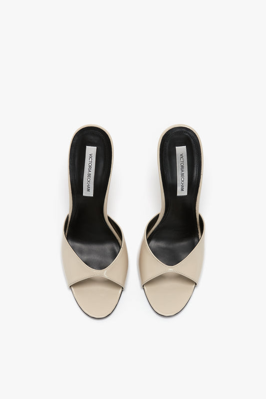 Top view of a pair of beige open-toe slide sandals with flattering toe cleavage and black insoles, crafted from luxury calf leather. The brand label reads "Classic Mule In Macadamia Calf Leather by Victoria Beckham" inside the heels.