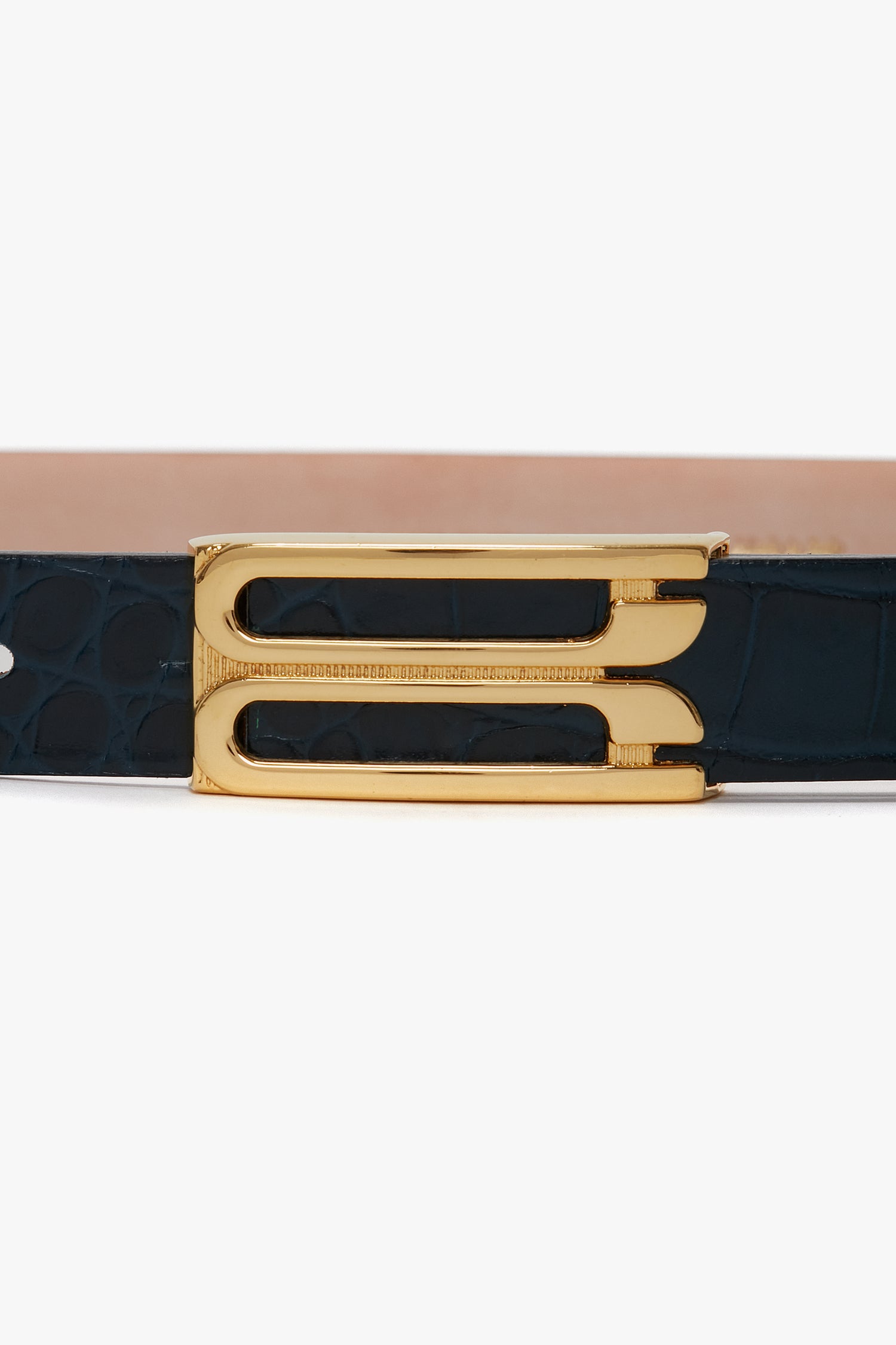 A close-up of a belt buckle attached to a Victoria Beckham Frame Belt In Midnight Blue Croc Embossed Calf Leather. The gold hardware buckle features a geometric design, and the belt has a light tan underside.