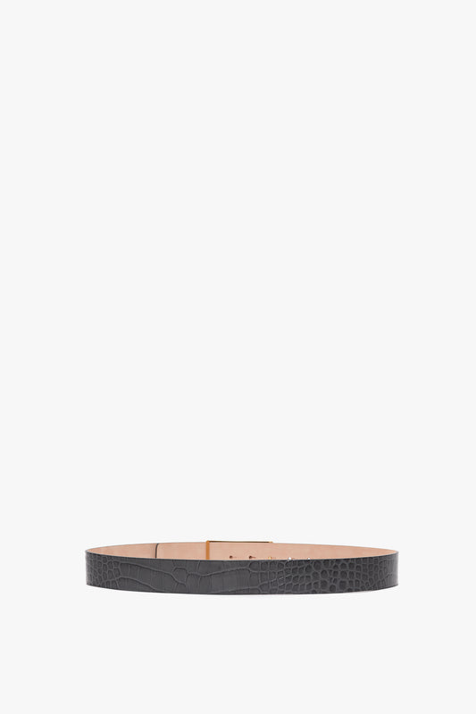 A black croc-embossed calf leather belt with a textured surface and tan interior is shown in a circular shape against a white background, featuring gold hardware for an added touch of elegance. This is the Jumbo Frame Belt In Slate Grey Croc Embossed Calf Leather by Victoria Beckham.