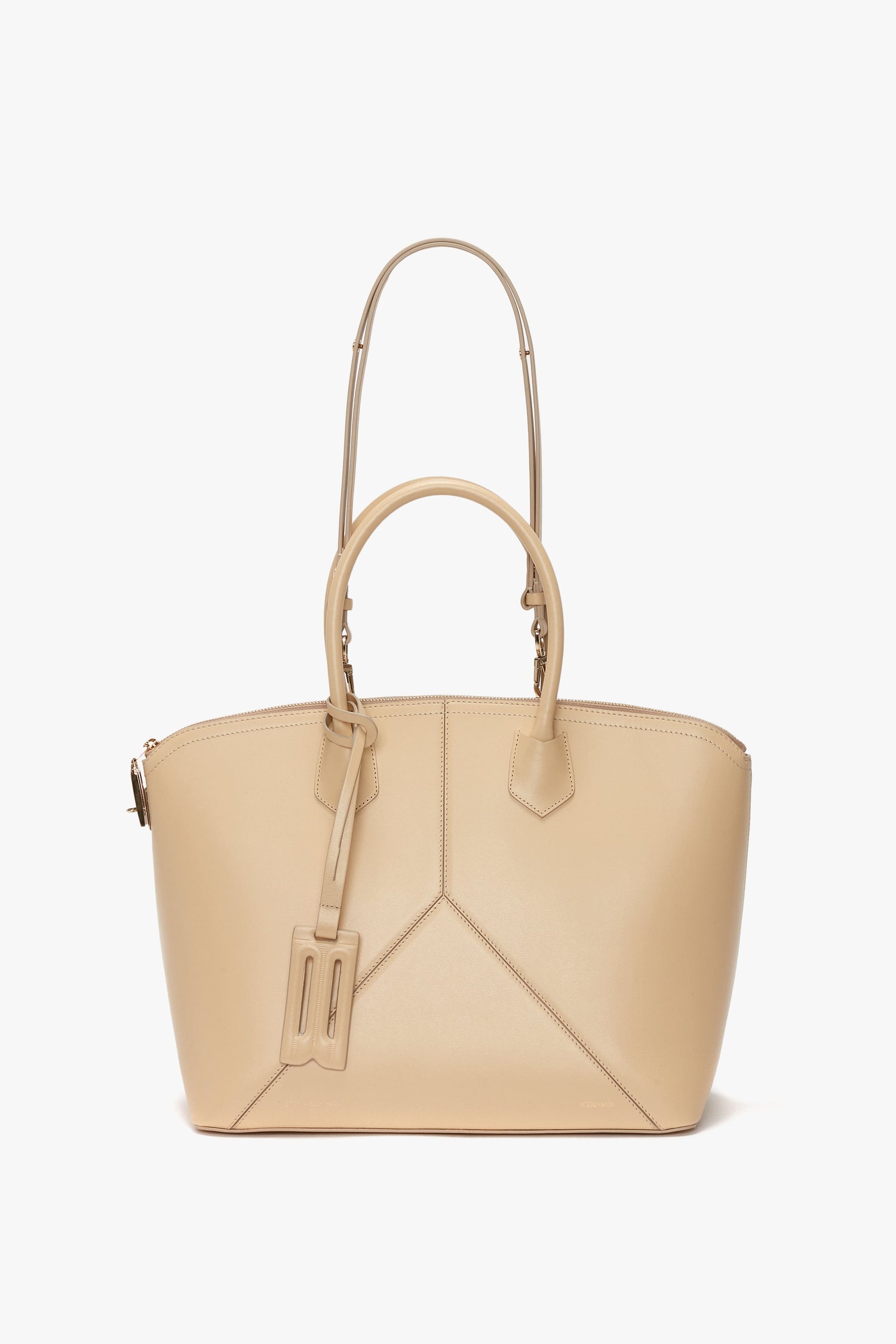 A tan Victoria Beckham Victoria Bag In Sesame Leather featuring dual handles, a detachable and adjustable shoulder strap, and elegant leather panels with a small tag attached.