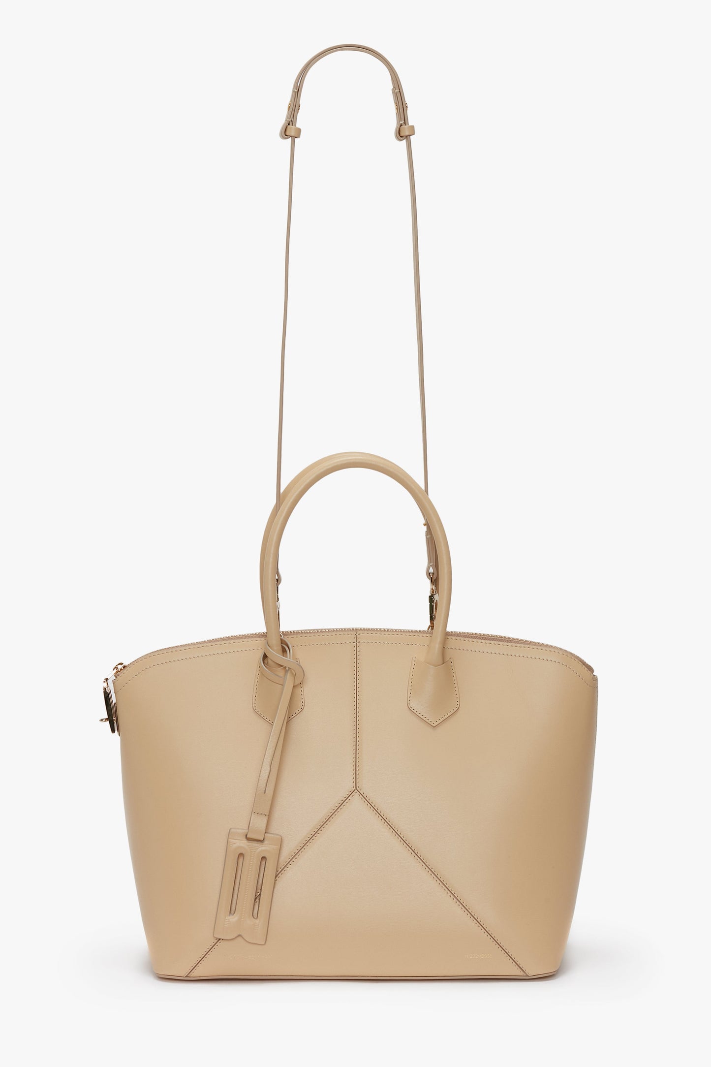 The Victoria Bag In Sesame Leather by Victoria Beckham is a beige leather handbag featuring leather panels, two short handles, and a decorative tag attached. It comes with a long adjustable strap and showcases a subtle geometric design on the front.