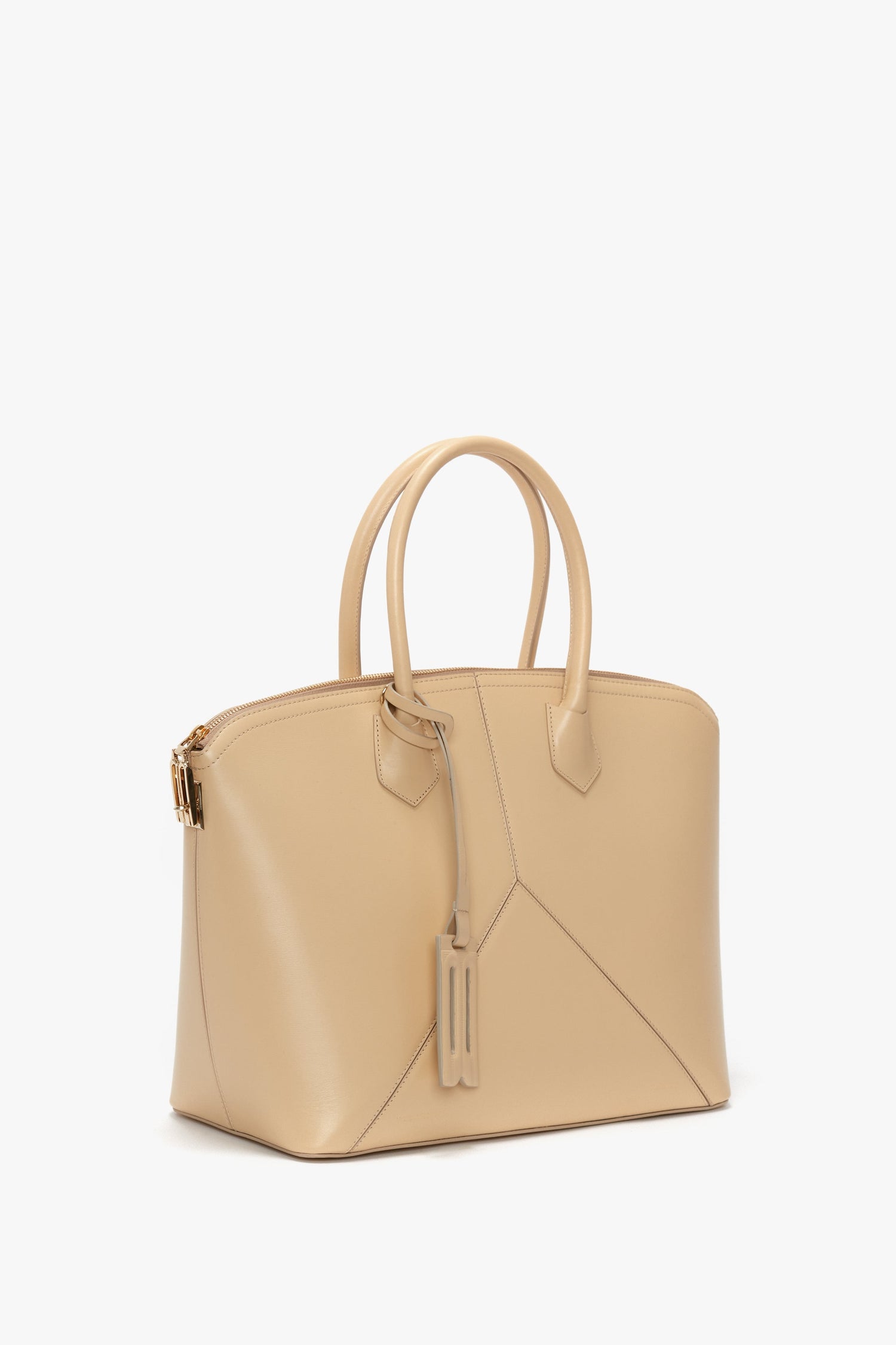 A tan leather Victoria Bag In Sesame Leather by Victoria Beckham featuring double handles, leather panels with a geometric design, and a hanging tag detail, all set against a white background.