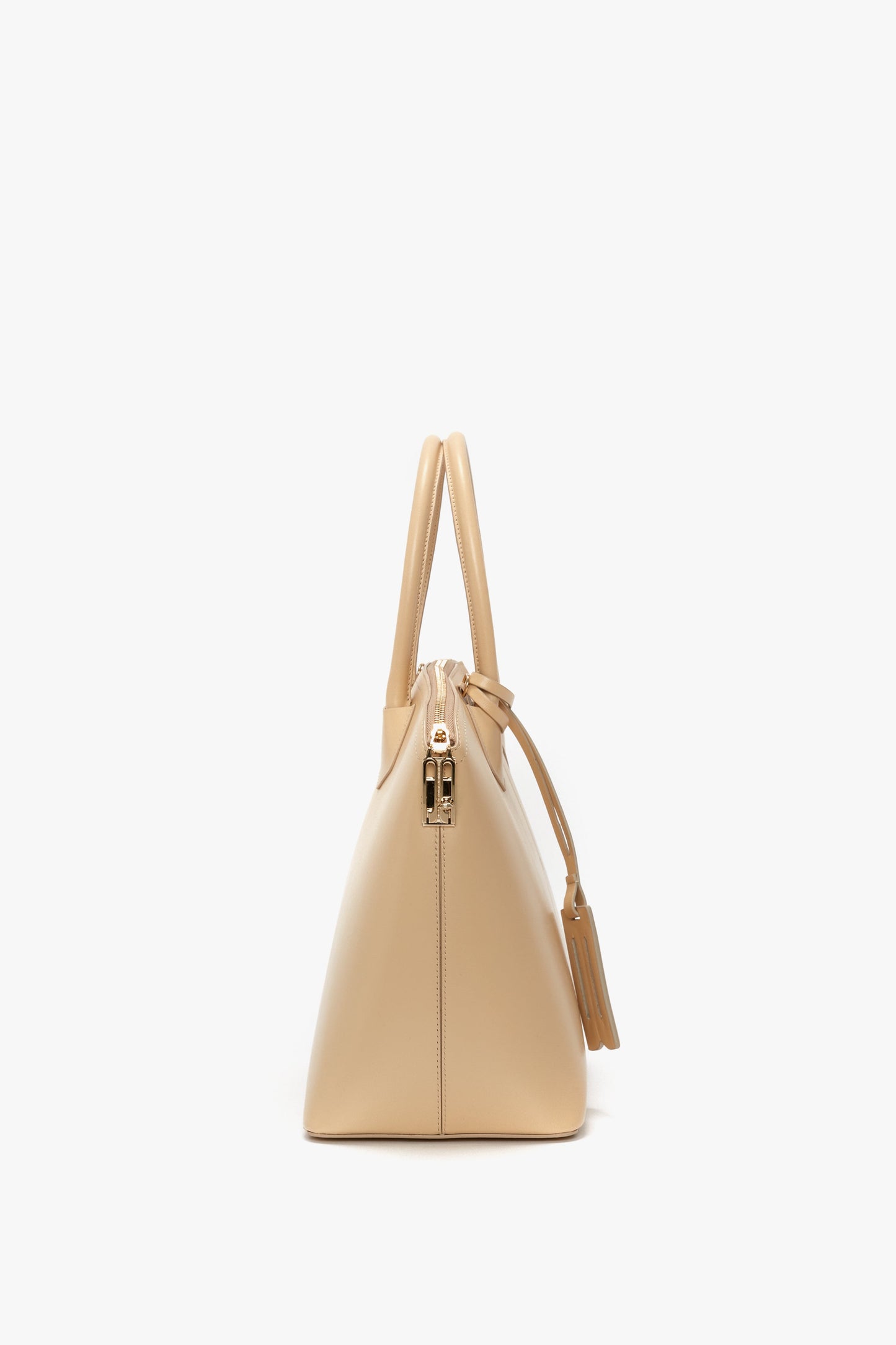Side profile of the Victoria Beckham Victoria Bag In Sesame Leather featuring beige leather panels, dual handles, a top zipper, and a keychain tassel on a white background.