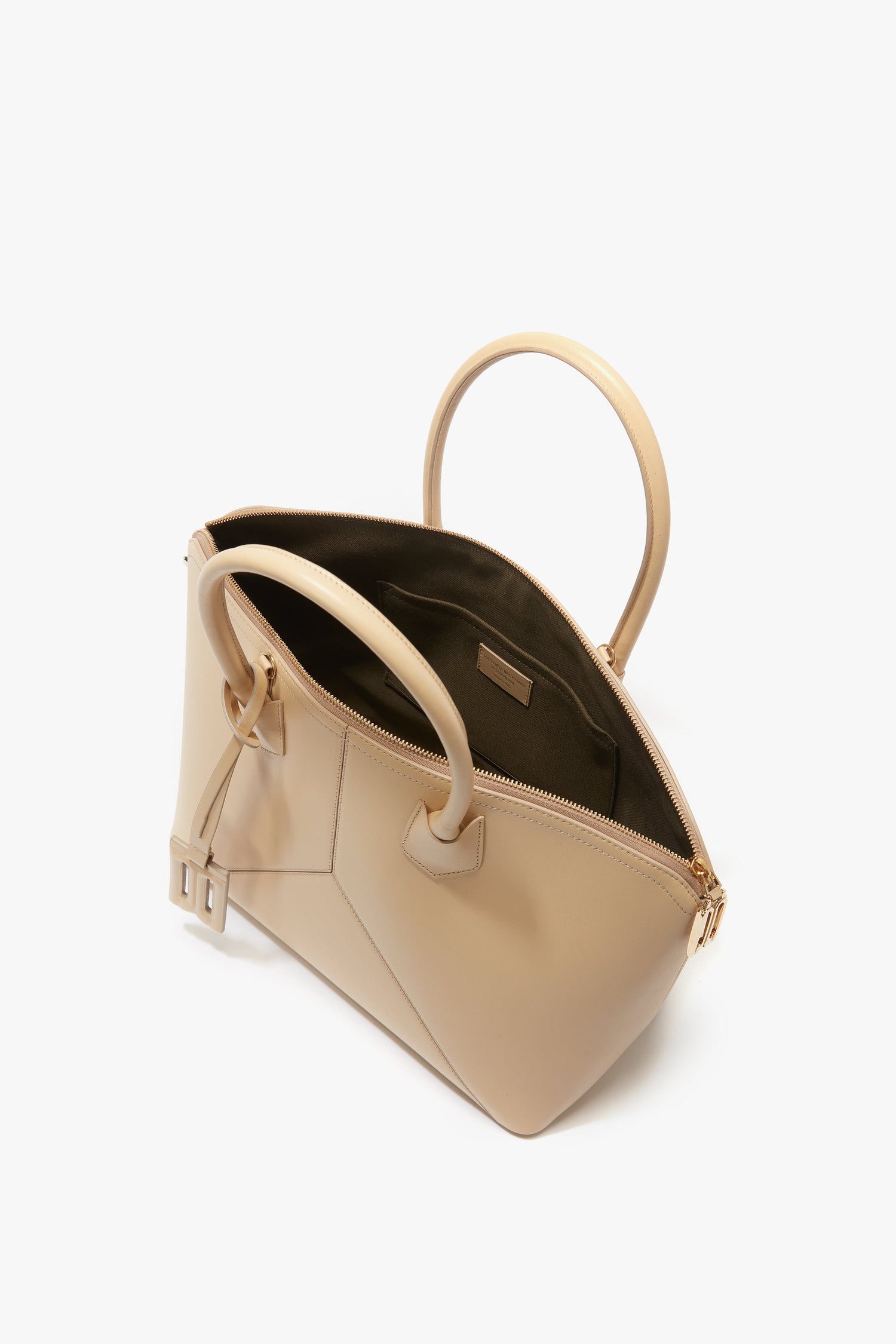 The Victoria Beckham Victoria Bag In Sesame Leather is a beige leather handbag with an open zippered top, two handles, leather panels, an interior pocket, and a small buckle detail on one side.