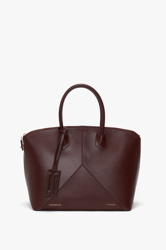 A Victoria Beckham Victoria Bag In Burgundy Leather with a structured design, featuring two handles, an adjustable strap, and a minimalistic tag.