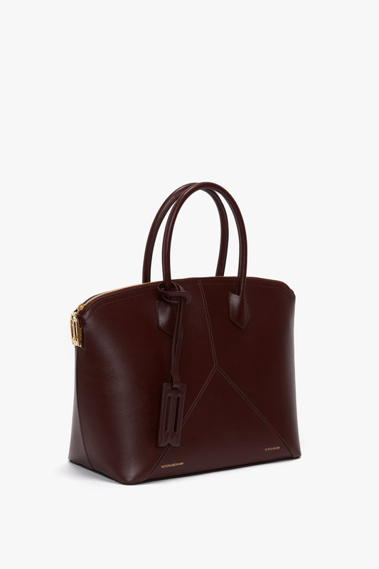 A dark brown Victoria Beckham Victoria Bag In Burgundy Leather with a structured design, double handles, an adjustable strap, a gold-toned zipper, and a small tag attached to one handle.