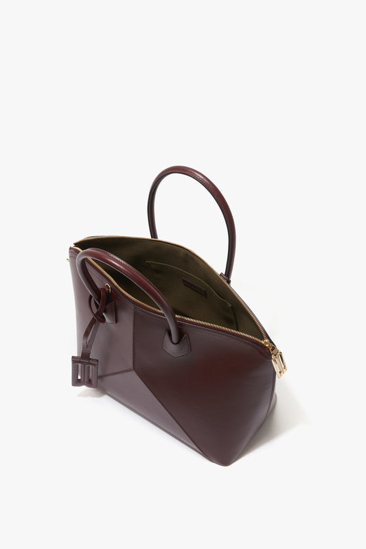 A brown leather handbag with a gold zipper, partially open to reveal a burgundy interior. Named the Victoria Bag In Burgundy Leather by Victoria Beckham, it features two handles, an adjustable strap, and a side tag.