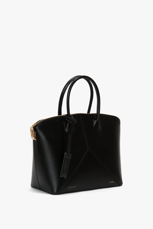A black leather tote handbag with dual handles, gold-tone hardware, and a rectangular tag. Featuring leather panels and a zippered top closure, the structured body is ideal for versatility. This Victoria Bag In Black Leather by Victoria Beckham effortlessly combines style with function.