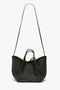 A black calf leather W11 Mini Tote Bag In Loden Leather by Victoria Beckham with long and short handles, featuring a minimalistic design and subtle branding at the bottom.