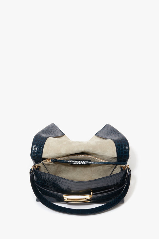 A B Pouch Bag In Croc Effect Midnight Blue Leather by Victoria Beckham is open, revealing a beige interior with a central zipped compartment and additional pockets.