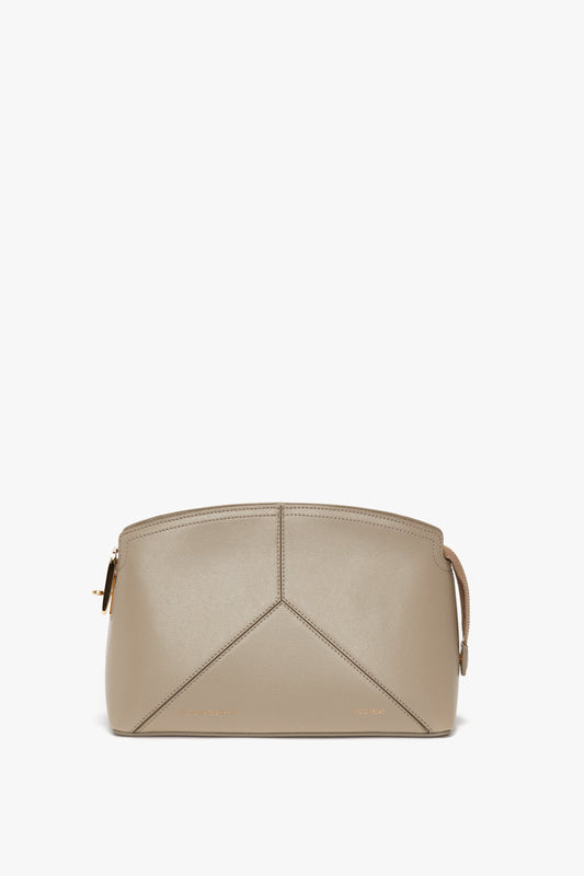 A taupe handbag with a minimalist design, crafted from premium calf leather and featuring a gold zipper, an adjustable and removable strap on the right, and subtle stitching details creating geometric shapes. The Victoria Crossbody Bag In Taupe Leather is part of the exquisite Victoria Beckham leather goods collection.