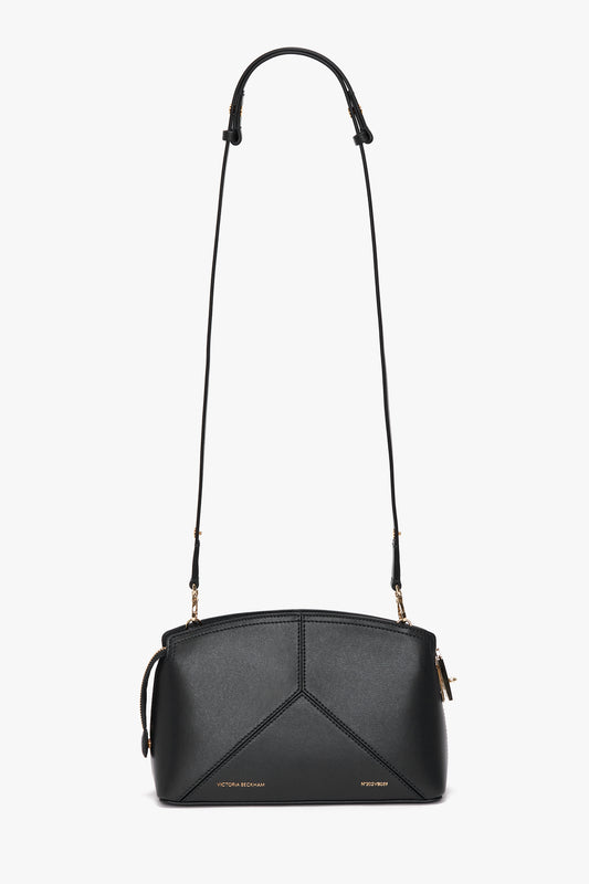 The Victoria Crossbody Bag In Black Leather is a black leather bag with an adjustable strap, gold-tone metal accents, and minimalistic stitching. Crafted from textured calf leather, it features the Victoria Beckham brand name embossed on the front.