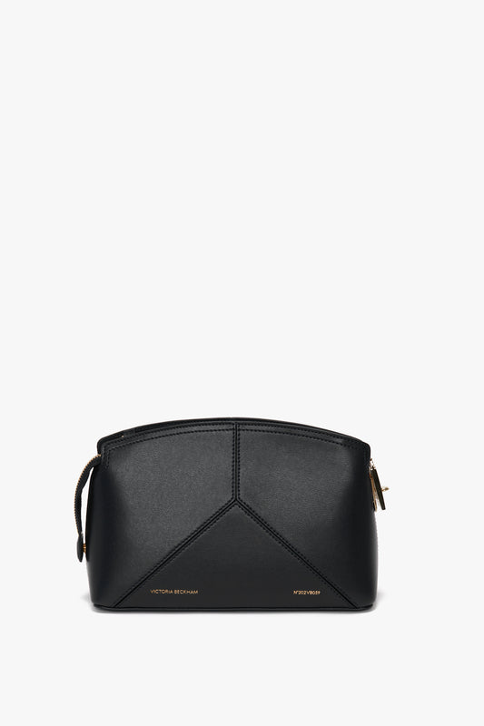A black, structured Victoria Crossbody Bag In Black Leather with gold-toned zipper and minimalistic geometric pattern. Made from textured calf leather, it features an adjustable strap for optimal comfort. "Victoria Beckham" and "Victoria Beckham" are stamped in gold on the front.