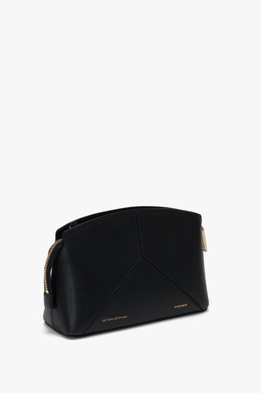 A black, structured handbag crafted from textured calf leather with gold zippers and "Victoria Beckham" embossed in gold at the bottom. The Victoria Crossbody Bag In Black Leather also features an adjustable strap for versatility.