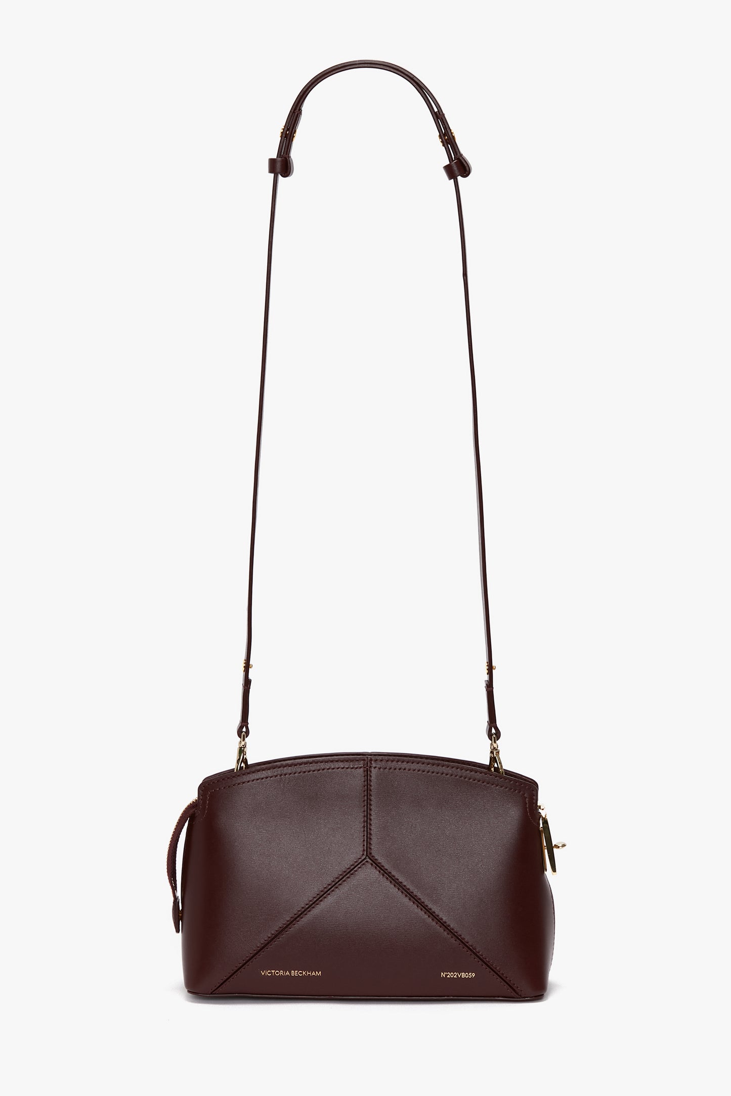 The Victoria Beckham Victoria Crossbody Bag In Burgundy Leather is a small, dark brown leather bag crafted from textured calf leather. It features a long adjustable strap, gold hardware, a padlock closure, and stitched geometric patterns on the front.