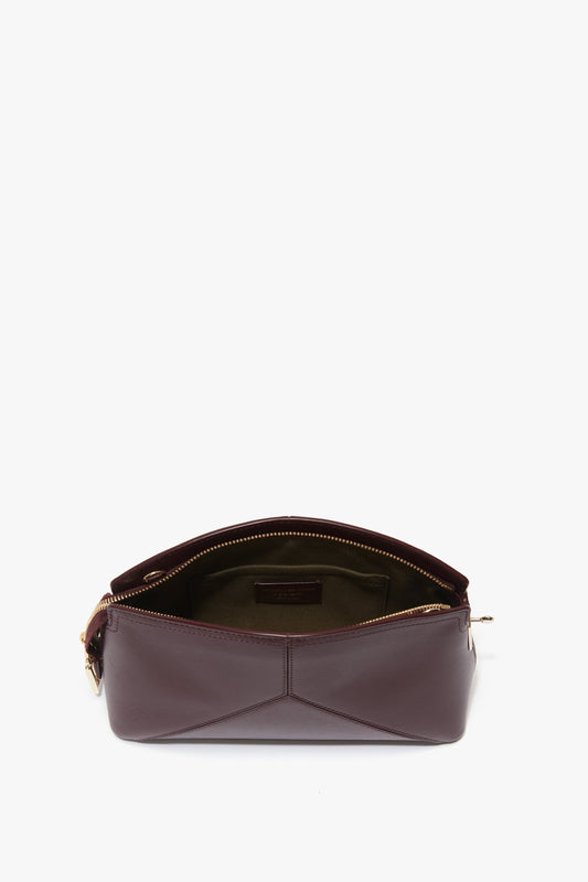 A maroon Victoria Crossbody Bag In Burgundy Leather by Victoria Beckham is open, showing a green interior with an internal pocket. The zipper is pulled back, revealing the contents and structure of the bag and its elegant padlock closure.