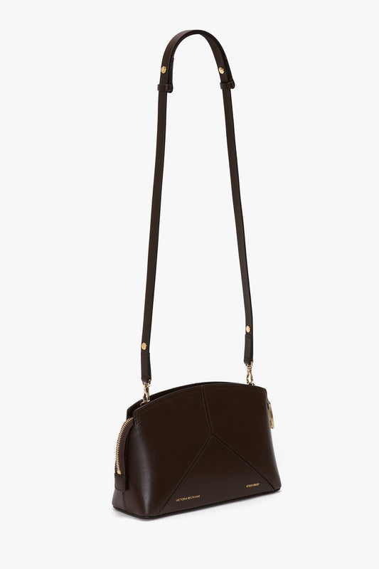 The Victoria Beckham Exclusive Victoria Crossbody Bag In Brown Leather is a small, dark brown shoulder bag made from premium calf leather, with a long adjustable strap and gold-colored hardware. It features a zippered top and a minimalist design, complete with the signature V shape.