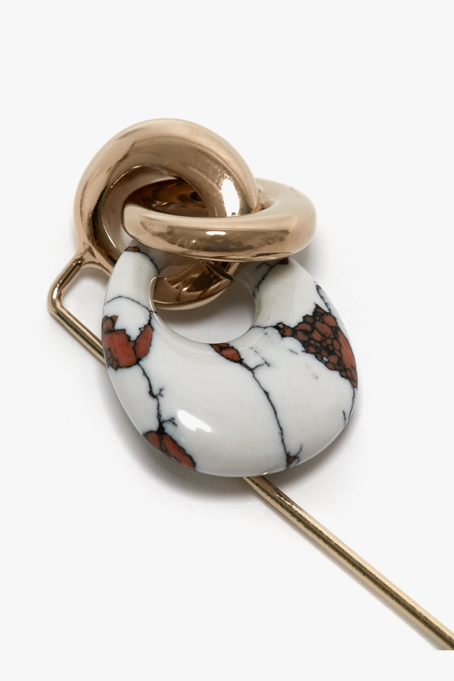 A Victoria Beckham Exclusive Resin Charm Brooch In Light Gold-White featuring an intricate design: a gold knot and a white marble-patterned ring with brown speckles, attached to a gold stick pin. This exquisite accessory, made in Italy, adds elegance to any look.
