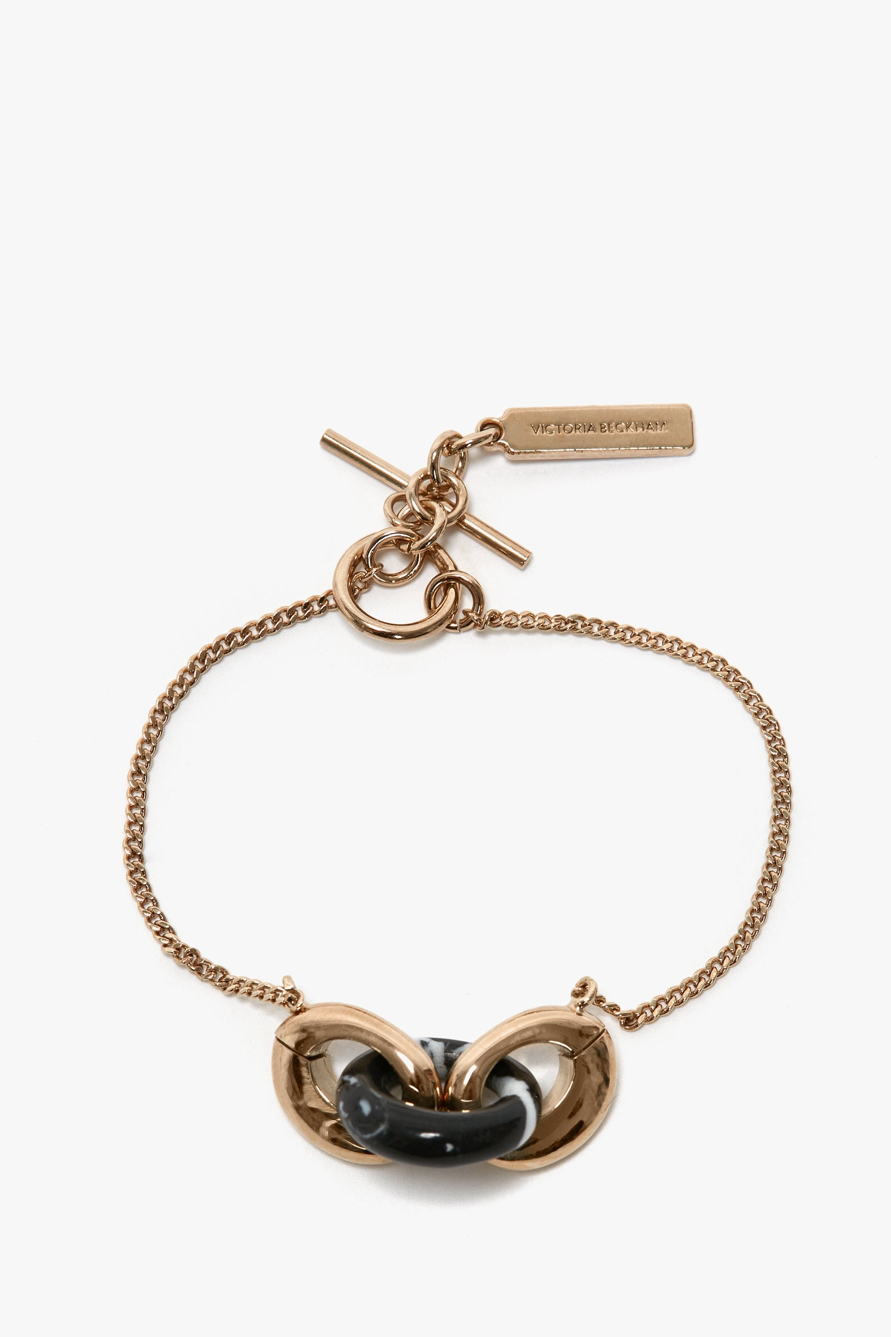 An Exclusive Resin Charm Bracelet In Light Gold-Black by Victoria Beckham, featuring a chain with a toggle clasp and an intertwined central design. Crafted from 100% brass, it includes a small rectangular tag attached to the clasp.