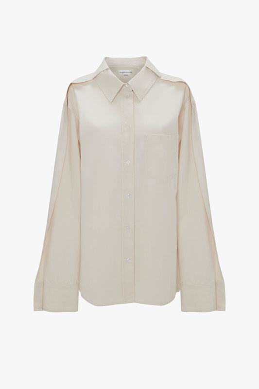A beige, oversized fit, long-sleeve button-up shirt with a collar and a single chest pocket displayed against a white background is the Victoria Beckham Oversized Pleat Detail Denim Shirt in Ecru.