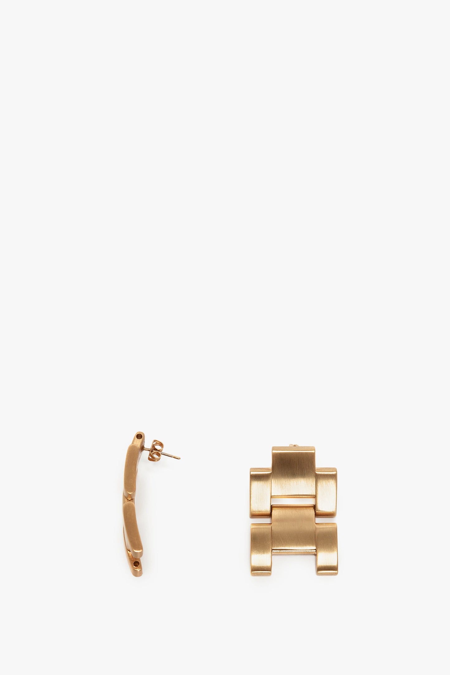 Two gold-toned metal earrings are shown. One earring is laid flat, and the other, featuring a Victoria Beckham logo, is displayed from its side to show the post and butterfly closing. The product name of these earrings is "Exclusive Jumbo Chain Earrings in Brushed Gold" by Victoria Beckham.