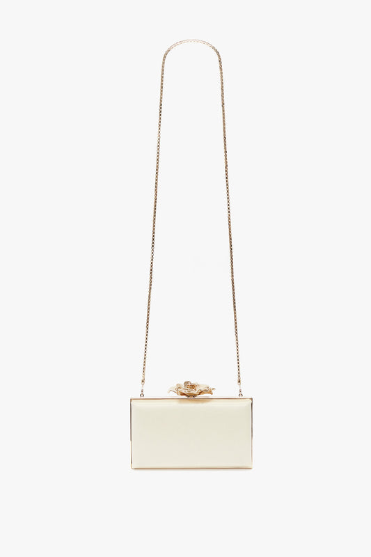 A rectangular white clutch purse with a bespoke brass frame and a long, thin gold chain strap, featuring a gold floral ornament on the top. Reminiscent of Victoria Beckham accessories, this Frame Flower Minaudiere in Chamomile by Victoria Beckham is both elegant and contemporary.
