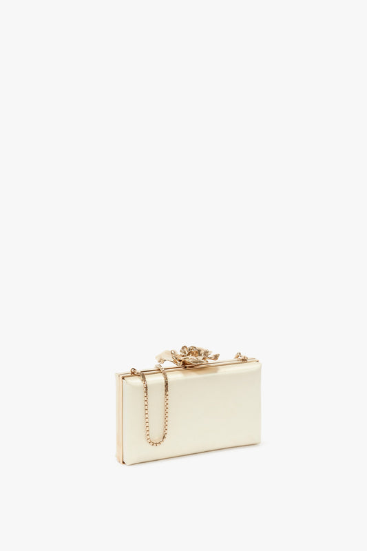 A cream-colored rectangular clutch with a bespoke brass frame, adorned with a gold chain strap and an ornate floral clasp on top, reminiscent of the Victoria Beckham Frame Flower Minaudiere in Chamomile.