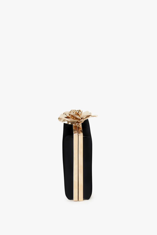A black and gold cylindrical gift box with a golden ribbon and bow on a white background, featuring a Victoria Beckham Frame Flower Minaudiere in Black.