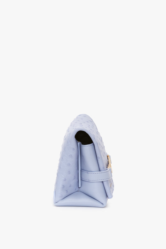 Side view of a pastel blue, textured Mini Chain Pouch With Long Strap In Frost Ostrich-Effect Leather by Victoria Beckham with a structured design, gold clasp, and an elegant crossbody bag strap.