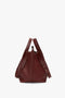 A W11 Medium Tote Bag In Burgundy Leather by Victoria Beckham with adjustable straps and gold-tone hardware, crafted from grained leather, stands upright on a plain white background.