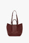 A W11 Medium Tote Bag In Burgundy Leather by Victoria Beckham, crafted from grained leather, featuring two short handles and an adjustable shoulder strap, displayed against a plain white background.