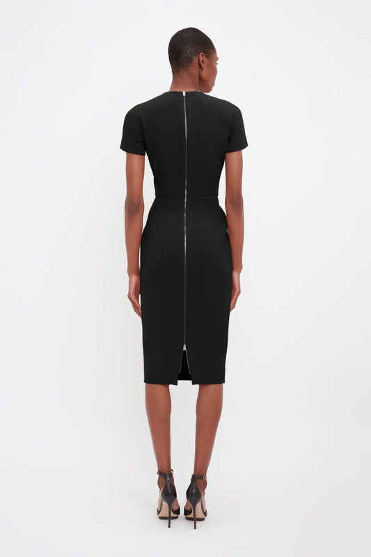 A woman standing in a studio, wearing a Victoria Beckham black knee-length fitted t-shirt dress made of matte bonded crepe with short sleeves and back zipper, viewed from behind.