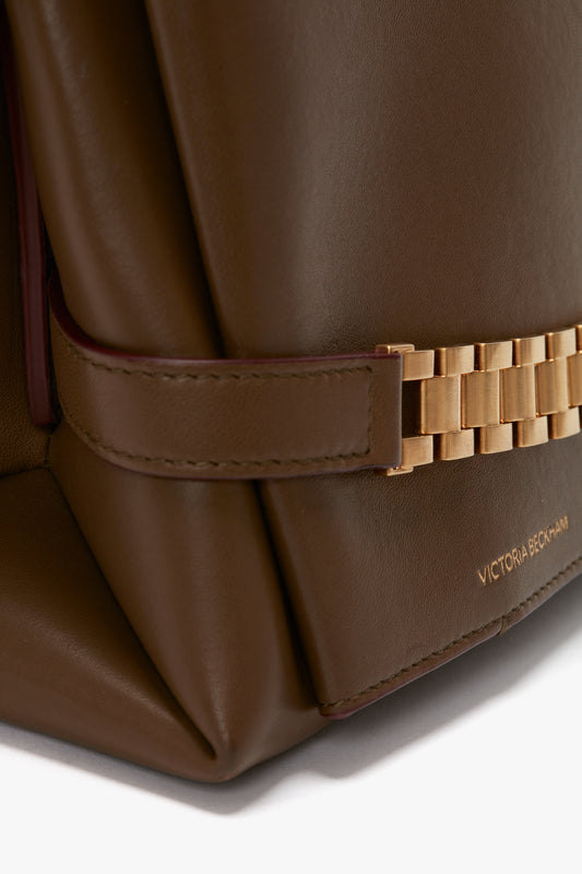 Close-up of a brown Nappa leather handbag featuring a gold chain detail and the name 'Victoria Beckham' embossed on the lower right corner. The stylish Chain Pouch Bag With Strap In Khaki Leather also includes a convenient detachable strap.