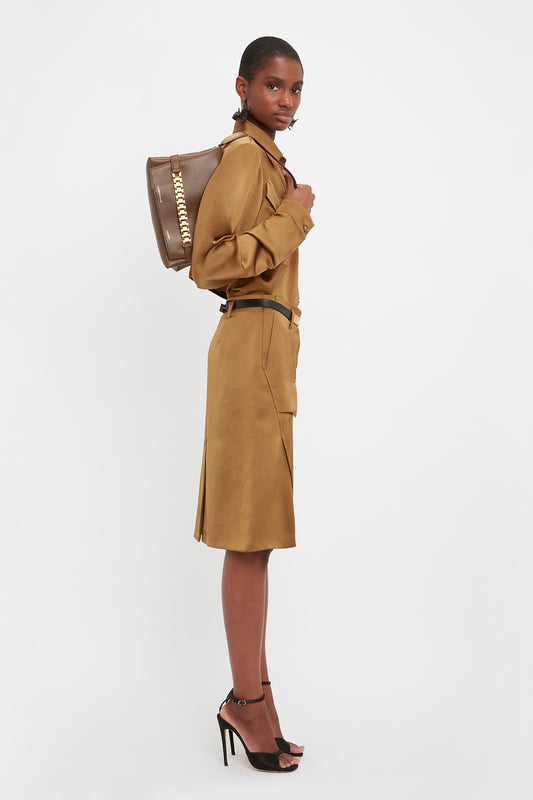 A person stands in profile, wearing a long-sleeved brown dress, black high heels, and carrying a Chain Pouch Bag With Strap In Khaki Leather by Victoria Beckham. The background is plain white.