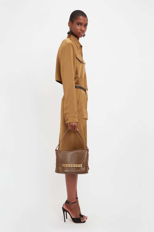 A person stands sideways wearing a long brown coat dress and black high heels, holding a Chain Pouch Bag With Strap In Khaki Leather from Victoria Beckham. The background is plain white.