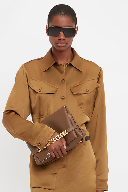 Person wearing oversized sunglasses and a brown button-up shirt, holding a Victoria Beckham Chain Pouch Bag With Strap In Khaki Leather.
