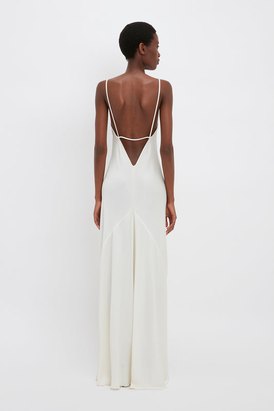 A person in a Victoria Beckham Floor-Length Cami Dress In Ivory, reminiscent of 90s fashion, stands facing away from the camera. The backless, floor-length dress features thin straps and a V-shaped back detail.