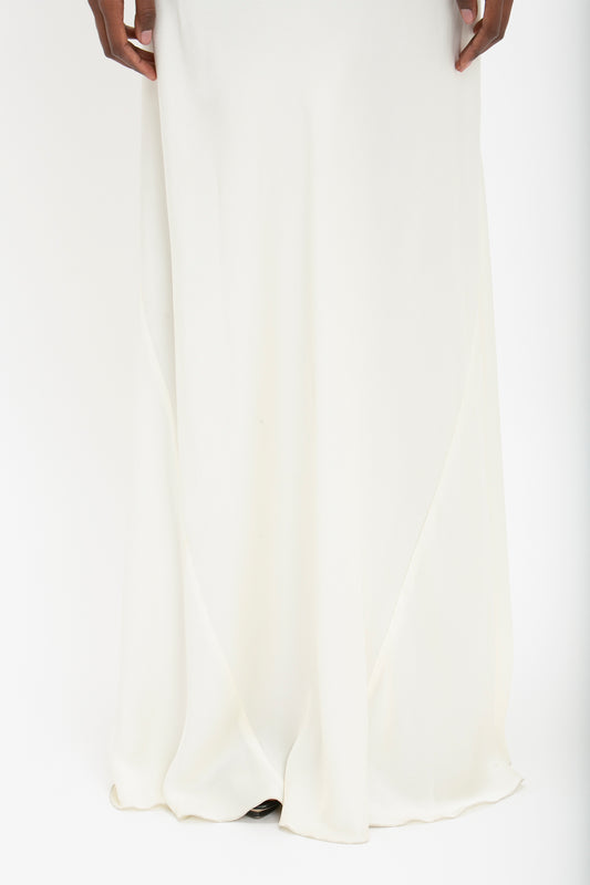 A person wearing a long, Floor-Length Cami Dress In Ivory by Victoria Beckham with both hands resting near the dress in front of a plain white background, exuding 90s fashion elegance.