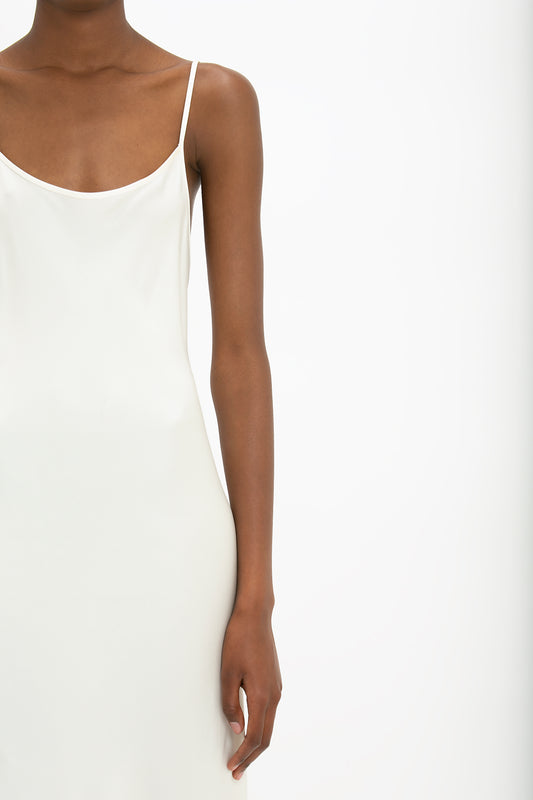 A person wearing a Victoria Beckham Floor-Length Cami Dress In Ivory stands against a plain background, evoking the effortless elegance of 90s fashion. Only the upper body from the shoulder to mid-thigh is visible, showcasing the sleek simplicity of crepe back satin.
