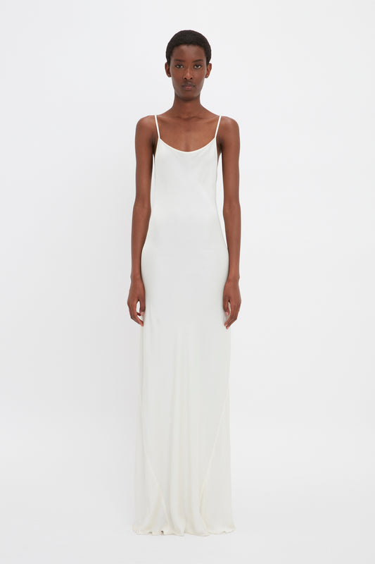 A person standing in a Victoria Beckham Floor-Length Cami Dress In Ivory with thin straps, reminiscent of 90s fashion. The background is plain white.