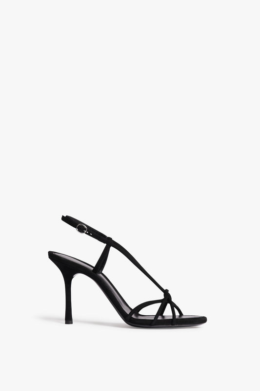 The Victoria Beckham Gaia Sandal in Black is a black high-heeled sandal with slim straps, an adjustable ankle strap, and a stiletto heel against a white background.