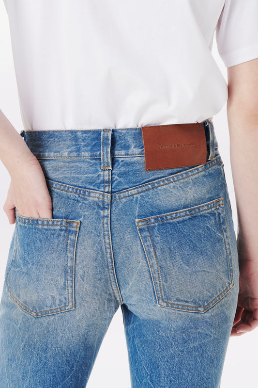 Back view of a person wearing a white shirt and a pair of Victoria Beckham's Victoria Mid-Rise Jean In Miami Wash with a brown leather patch on the waistband displaying the brand name. The mid-rise jeans, featuring a button fly, have one hand casually tucked in the back pocket.