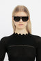 A young woman wearing Victoria Beckham's V Plaque Frame Sunglasses In Black and a black textured top, standing against a white background.