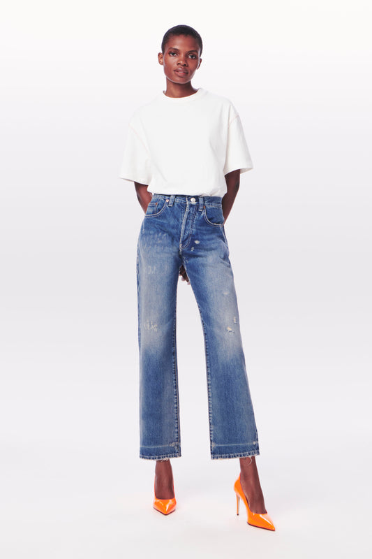 A person stands wearing a white t-shirt made of 100% cotton, Victoria Mid-Rise Jean in Vintage Wash by Victoria Beckham, and orange high-heeled shoes against a light grey background.