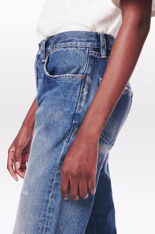 A person wearing a white top and Victoria Mid-Rise Jean in Vintage Wash by Victoria Beckham stands against a plain background. Their left arm is visible, hanging straight down by their side.