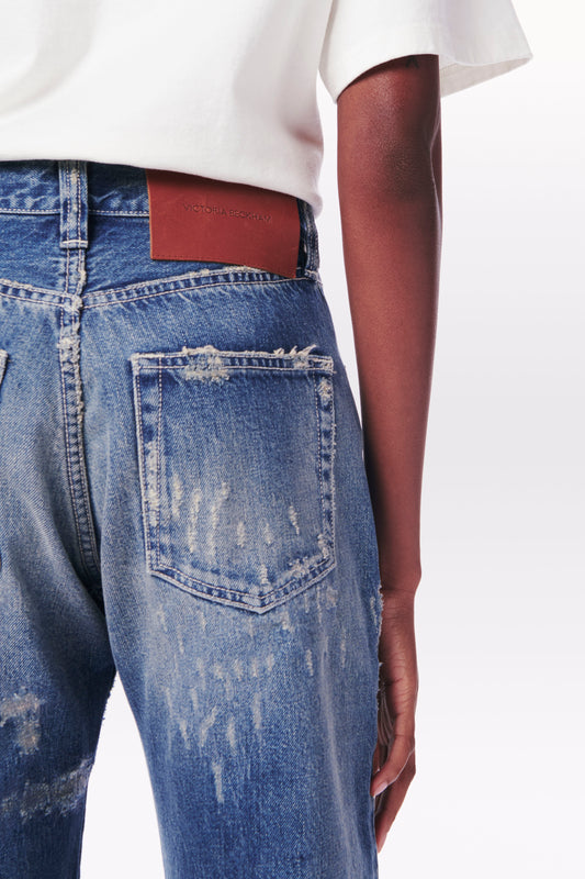 Close-up of a person wearing Victoria Beckham's Victoria Mid-Rise Jean in Vintage Wash with a distressed look, featuring a back pocket and brown leather patch on the waistband, paired with a white 100% cotton t-shirt.