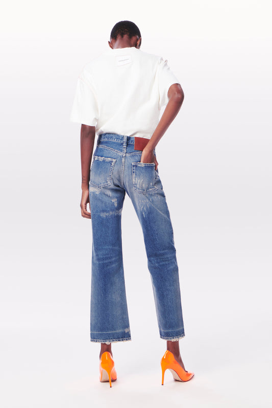A person with short hair is standing with their back to the camera, wearing a white shirt, Victoria Mid-Rise Jean in Vintage Wash by Victoria Beckham, and high-heeled orange shoes against a plain white background.