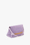 A Victoria Beckham Chain Pouch Bag with Strap in Lilac Suede with a light pink detachable strap and gold chain embellishment on the front. The Italian leather bag has a triangular shape and rests elegantly on a white surface.